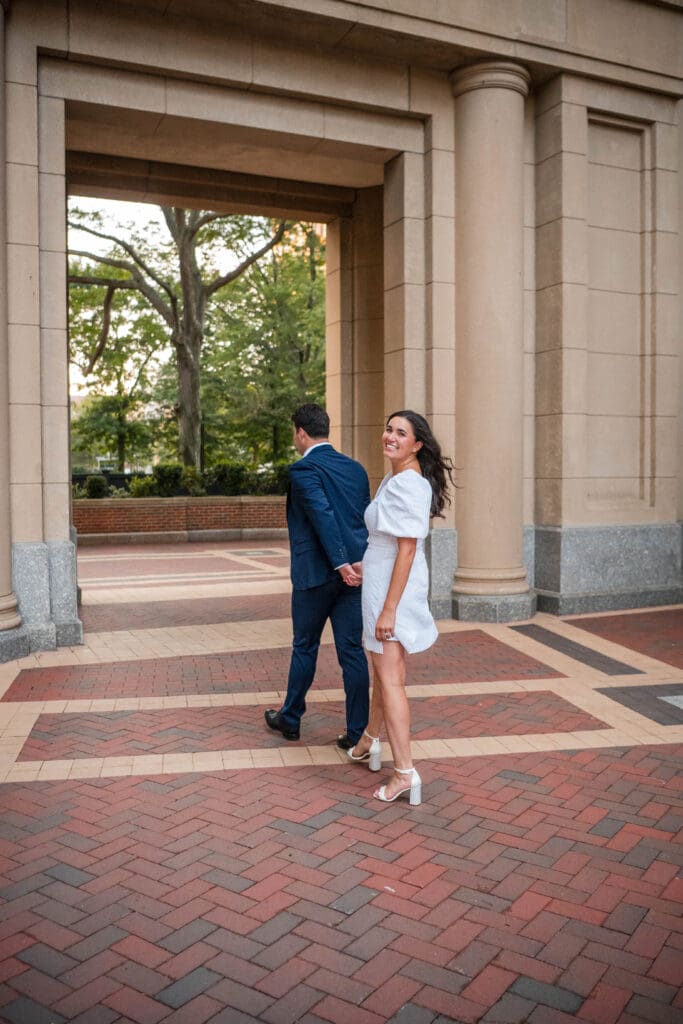 Man leads women as she looks back at the camera near the Harborwalk in Boston for an engagement photo.