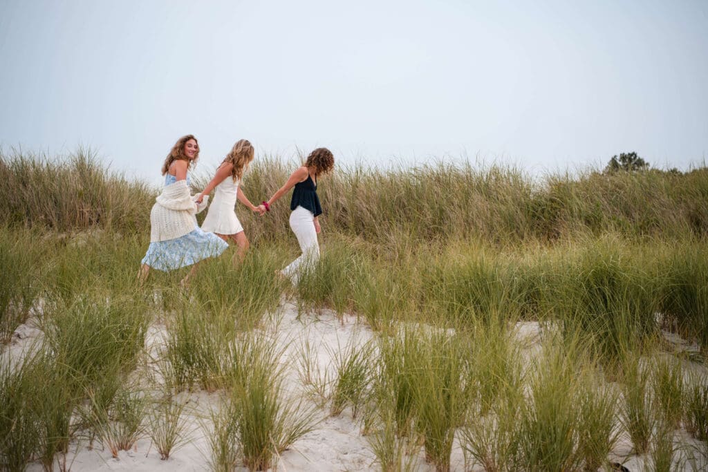 Three sisters walk hand in hand along the banks of Wingaersheek Beach. The last sister has a sweater slumped down her shoulders and looks at the camera.
