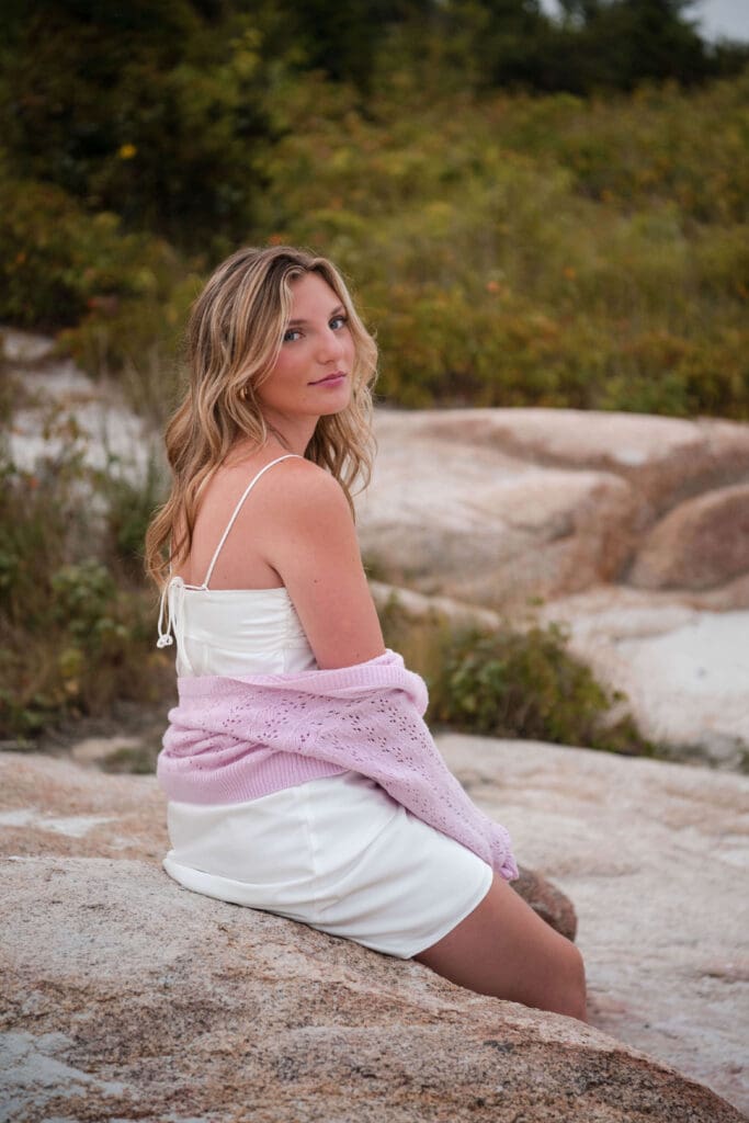 A senior Sits on a rock at Wingaersheek beach and gives a sift smile over her shoulder at the camera with her pink cardigan sweater halfway down her arms.