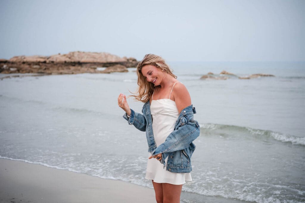 A girl standing on Wingaersheek Beach in her dress with her jean jacket halfway off her shoulders as she laughs with the wind blowing her hair. 