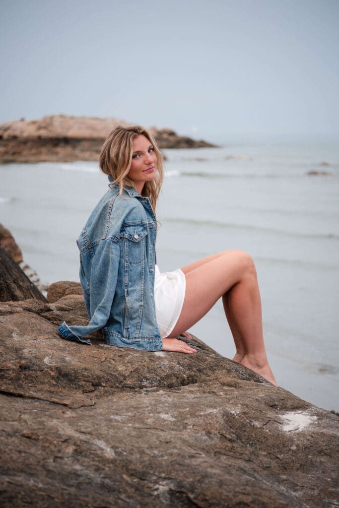 A girl sits facing the ocean at Wingaersheek Beach in a dress with her jean jacket draped over her shoulders and smiles back at the camera. 