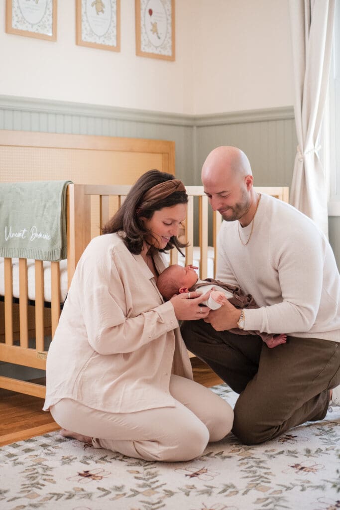 A Wakefield newborn photographer captures a couple kneeling down in front of the crib and smiling at their baby in this Nor Shore newborn session.
